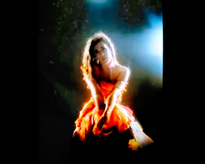 Imogen Heap in a music video with dramatic lighting, wearing an orange dress.